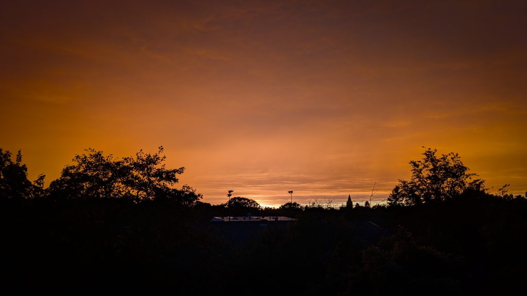 Orange leuchtender Himmel über eine schwarze Silhouette von Bäumen und Gebäuden.